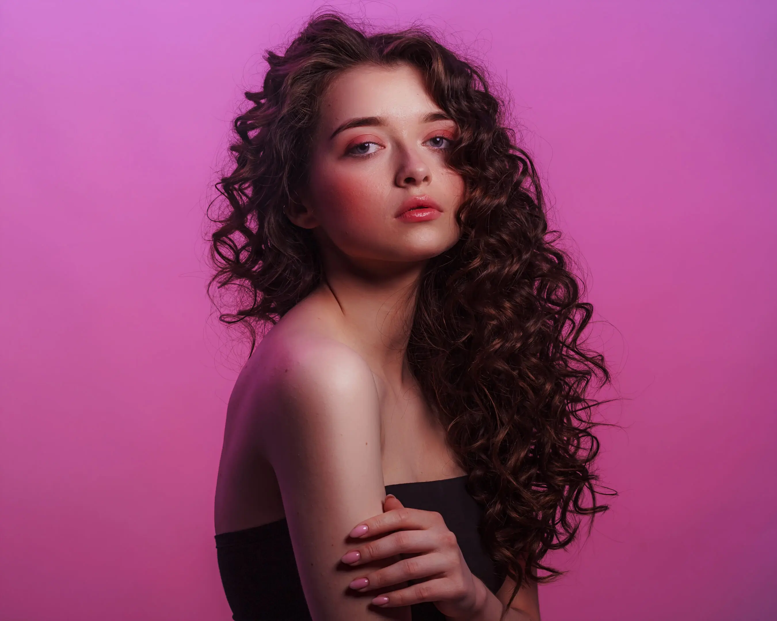 A person with long, curly brown hair poses against a pink background, flaunting the results of waxing in a strapless black top, with their hand gently touching one shoulder.