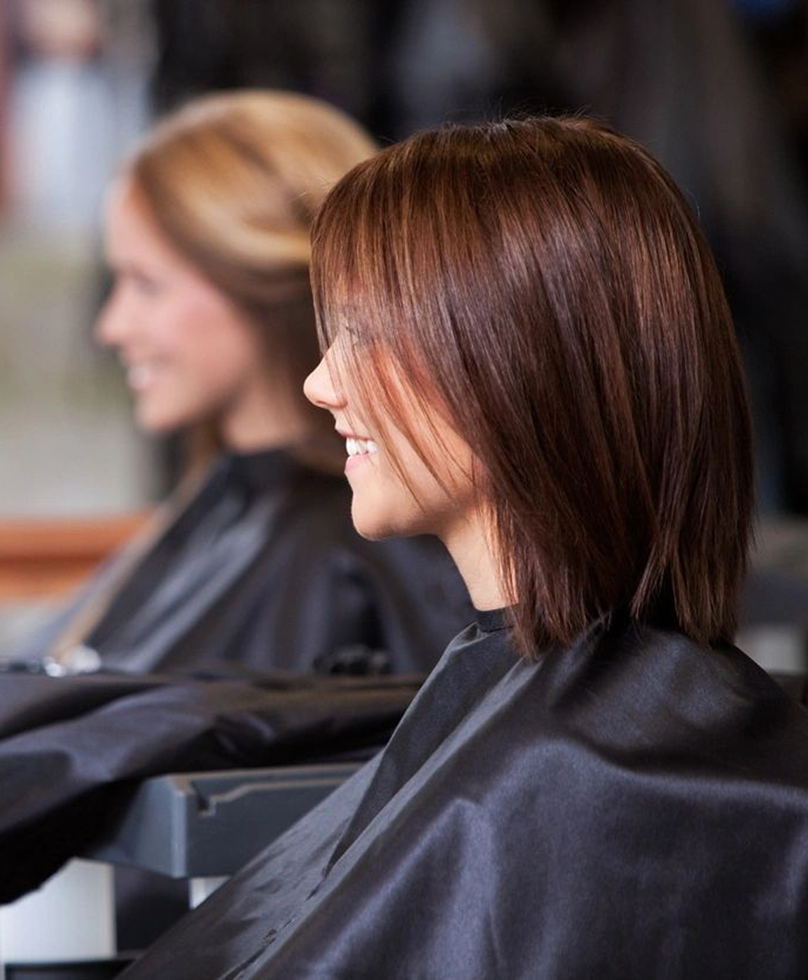 Two people with shoulder-length hair are seated in a salon, wearing black capes. They are turned slightly to the side, facing mirrors, as they consult with stylists about hair color services.
