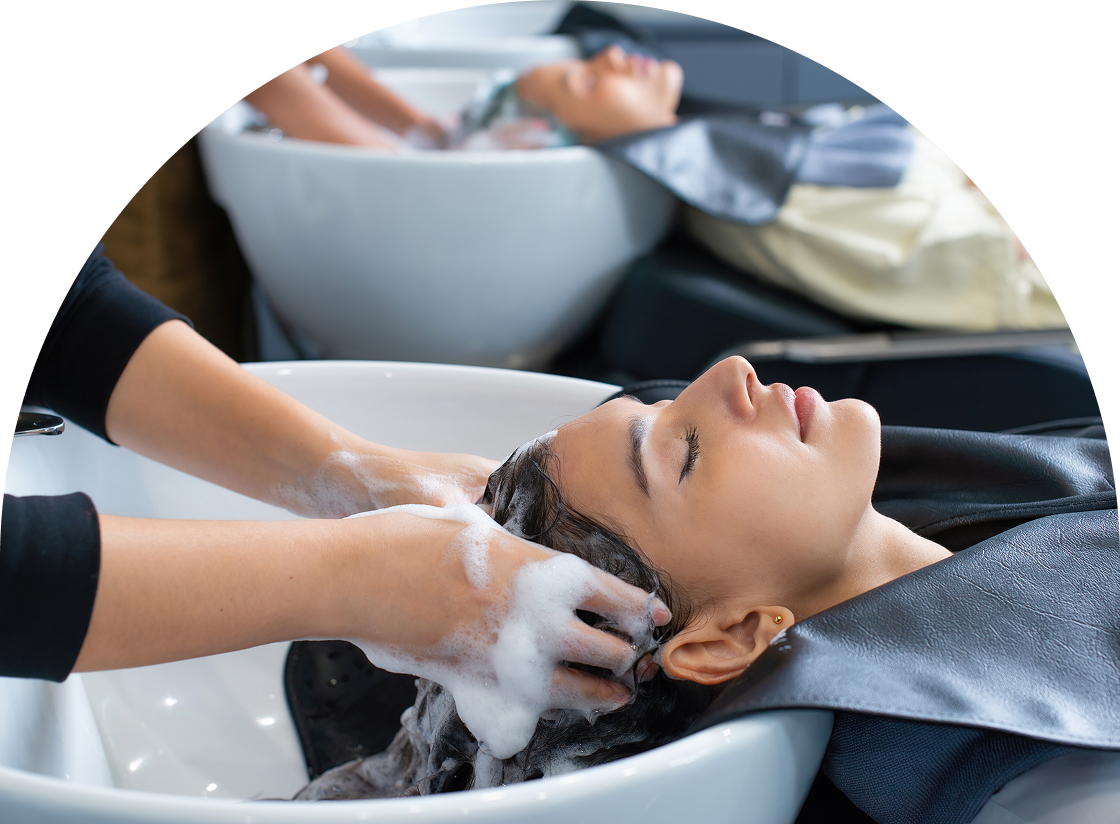 A person relaxes at the salon, eyes closed as their hair is gently washed in preparation for expert Hair Color Services. In the background, another client enjoys the same luxurious treatment.