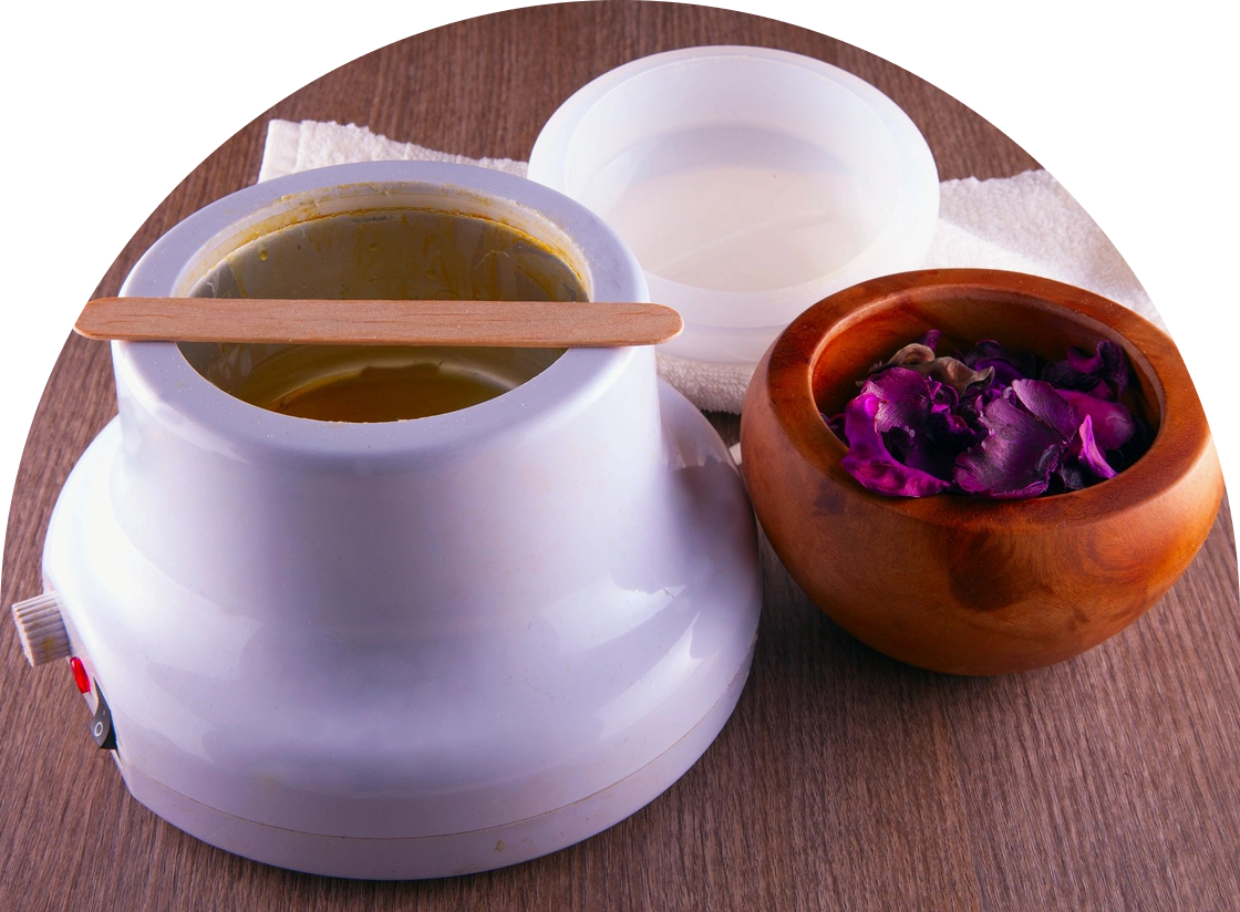 A wax warmer with a wooden stick, perfect for waxing and hair removal sessions, sits on a table next to a bowl of dried rose petals.