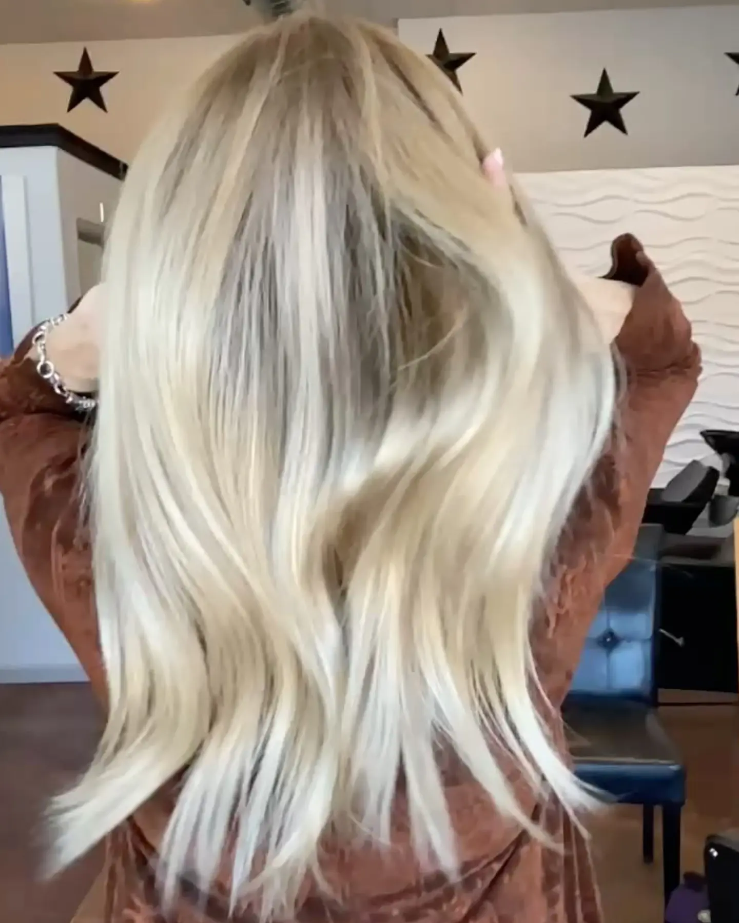 A person with long, blonde hair is shown from the back in a professional salon setting, holding their hair up with their hands.