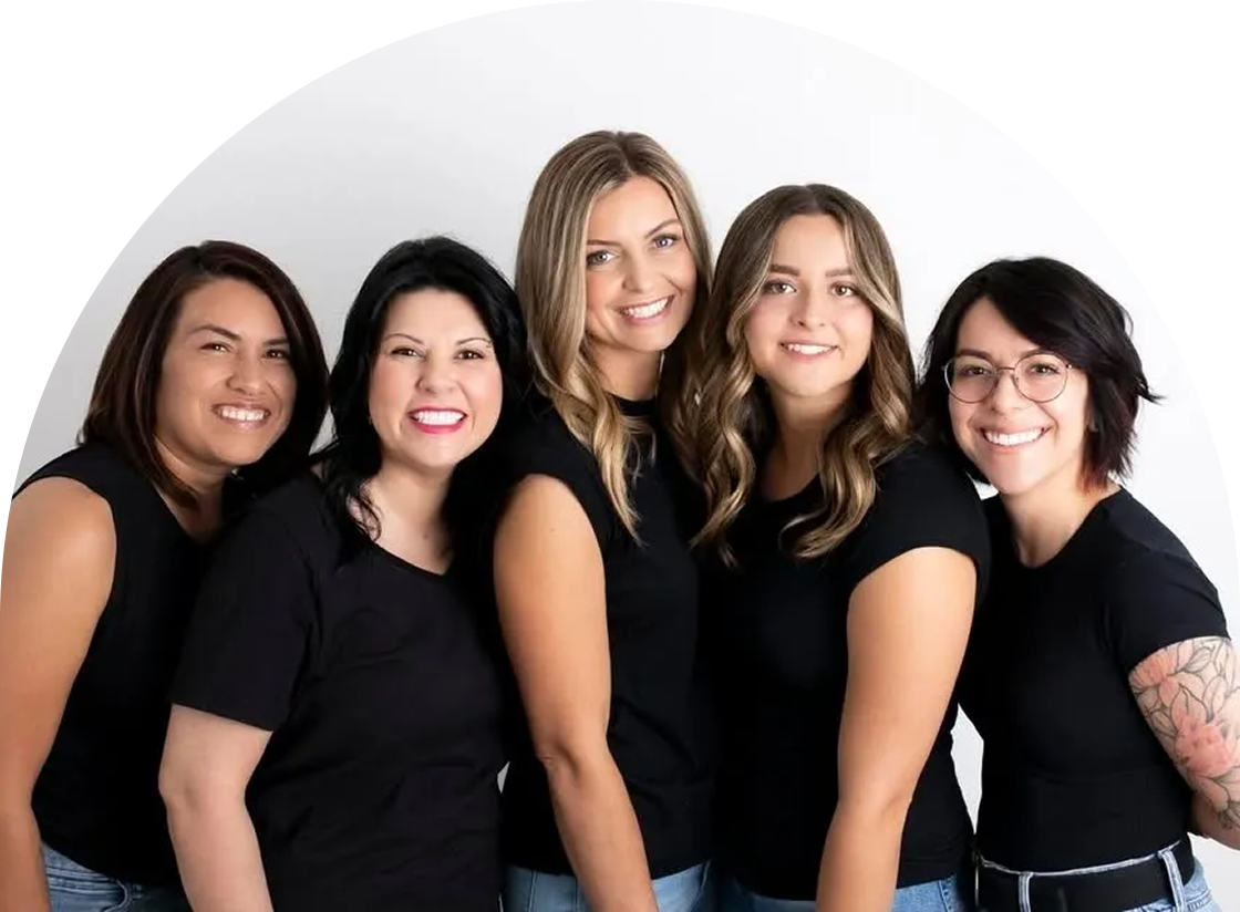 Five smiling people, showcasing their diverse hair color services, stand closely together in matching black shirts and jeans against a crisp white background.